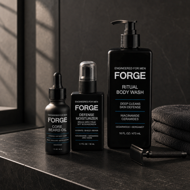The FORGE Full Line on a dark stone bathroom counter in warm morning window light: Core Beard Oil, Defense Moisturizer SPF 30 and Ritual Body Wash beside a black safety razor and charcoal towels